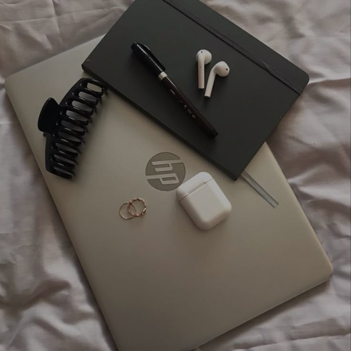 HP laptop with black hair clip, black notebook, black pen, wireless earbuds, and hoop earrings on white fabric