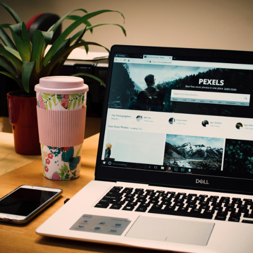 Dell laptop displaying Pexels website on wooden desk with colorful reusable coffee cup and smartphone