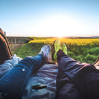 Two people relaxing in a car trunk with legs stretched out, overlooking a sunset and yellow flower field