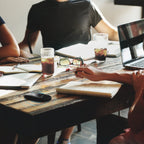 People working at a rustic wooden table with laptops, notebooks, drinks, and sunglasses in a casual meeting