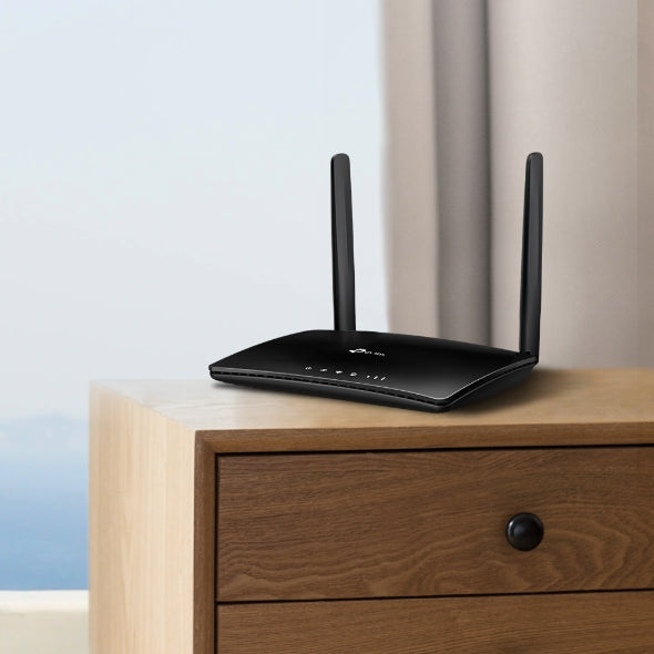 Black dual-antenna WiFi router placed on wooden dresser near window and curtains